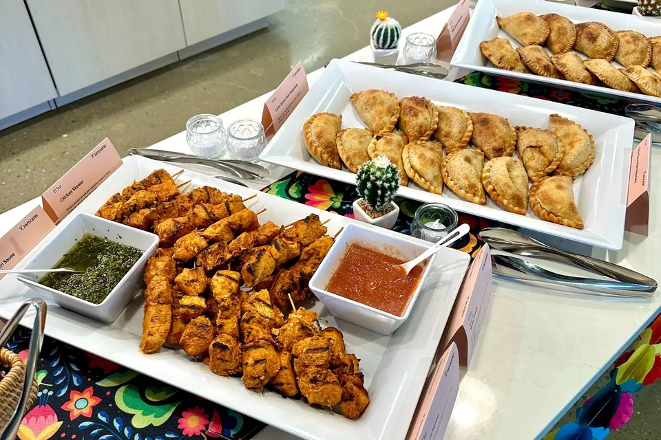 Office catering table with white platters of empanadas and chicken skewers, each platter fronted by a printed card naming the dish