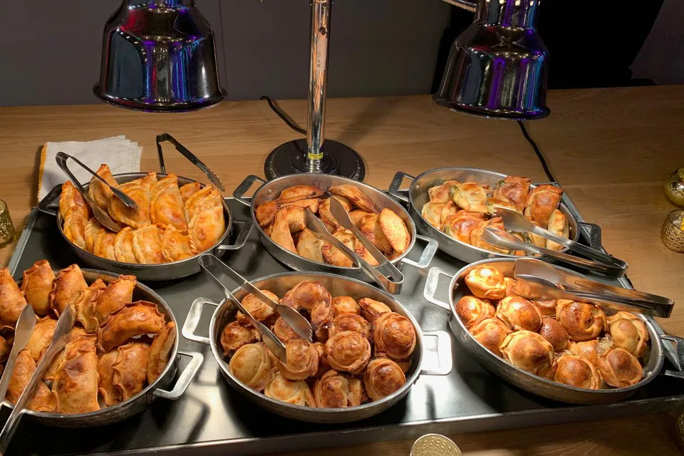 Six metal warming pans of baked empanadas under heat lamps on a wooden office counter, each pan with its own serving tongs