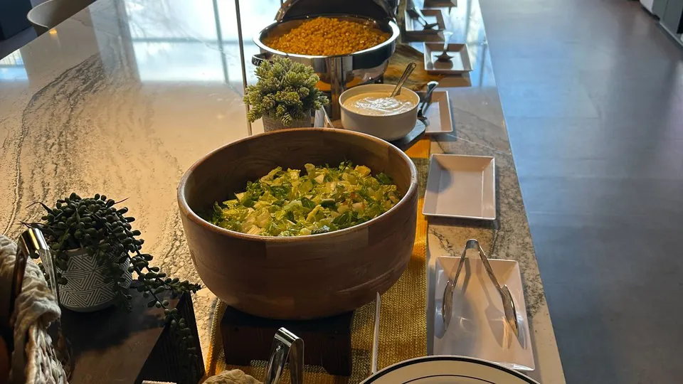 Office buffet on a marble counter with a wooden bowl of chopped salad, a chafing dish of dal, a bread basket and a stack of plates