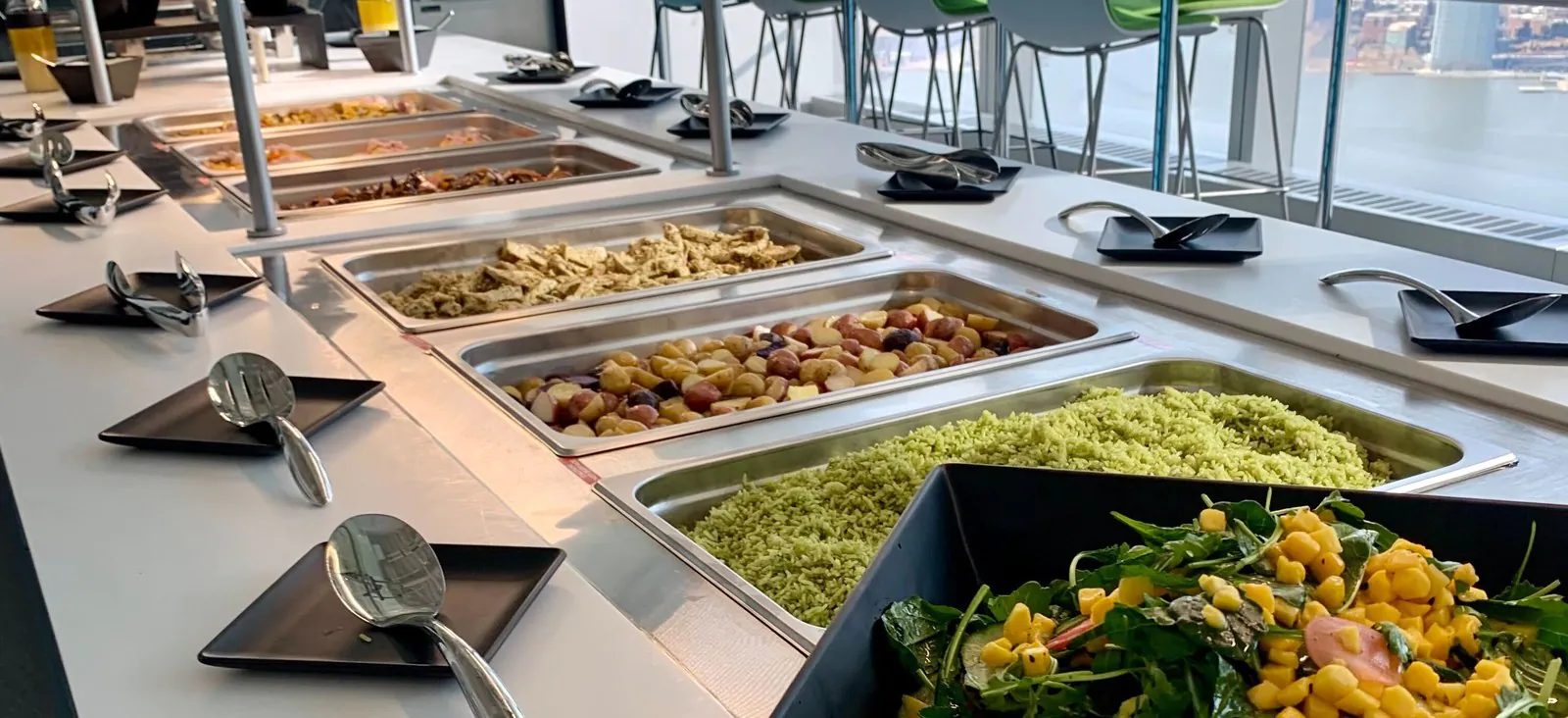 Office cafeteria serving line with stainless hotel pans of chicken, roasted potatoes and herbed rice behind a sneeze guard, city skyline through the window