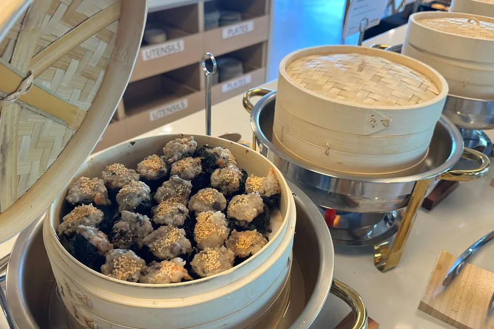 An open bamboo steamer of sesame-topped bites on an office buffet line, with further steamers on chafing stands behind