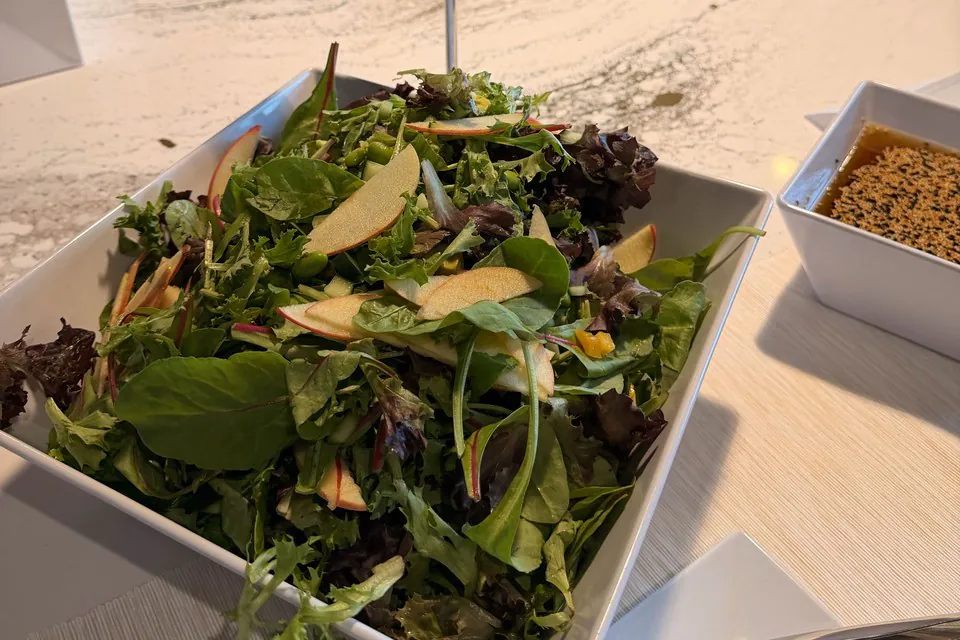 A large square bowl of mixed greens with apple slices beside a ramekin of sesame dressing on an office counter