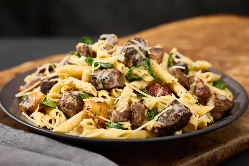 A plate of penne with seared steak pieces, mushrooms, spinach, blistered tomatoes and grated Parmesan on a wooden board
