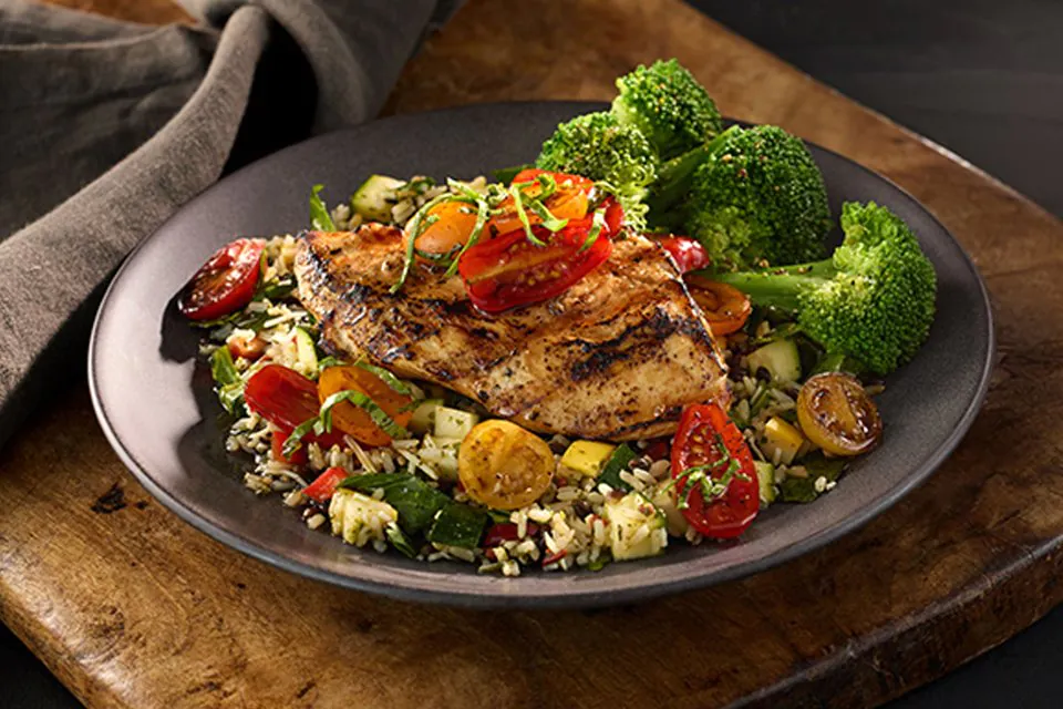 A grilled chicken breast topped with glazed cherry tomatoes and basil on a bed of grains and vegetables, with broccoli, on a dark plate