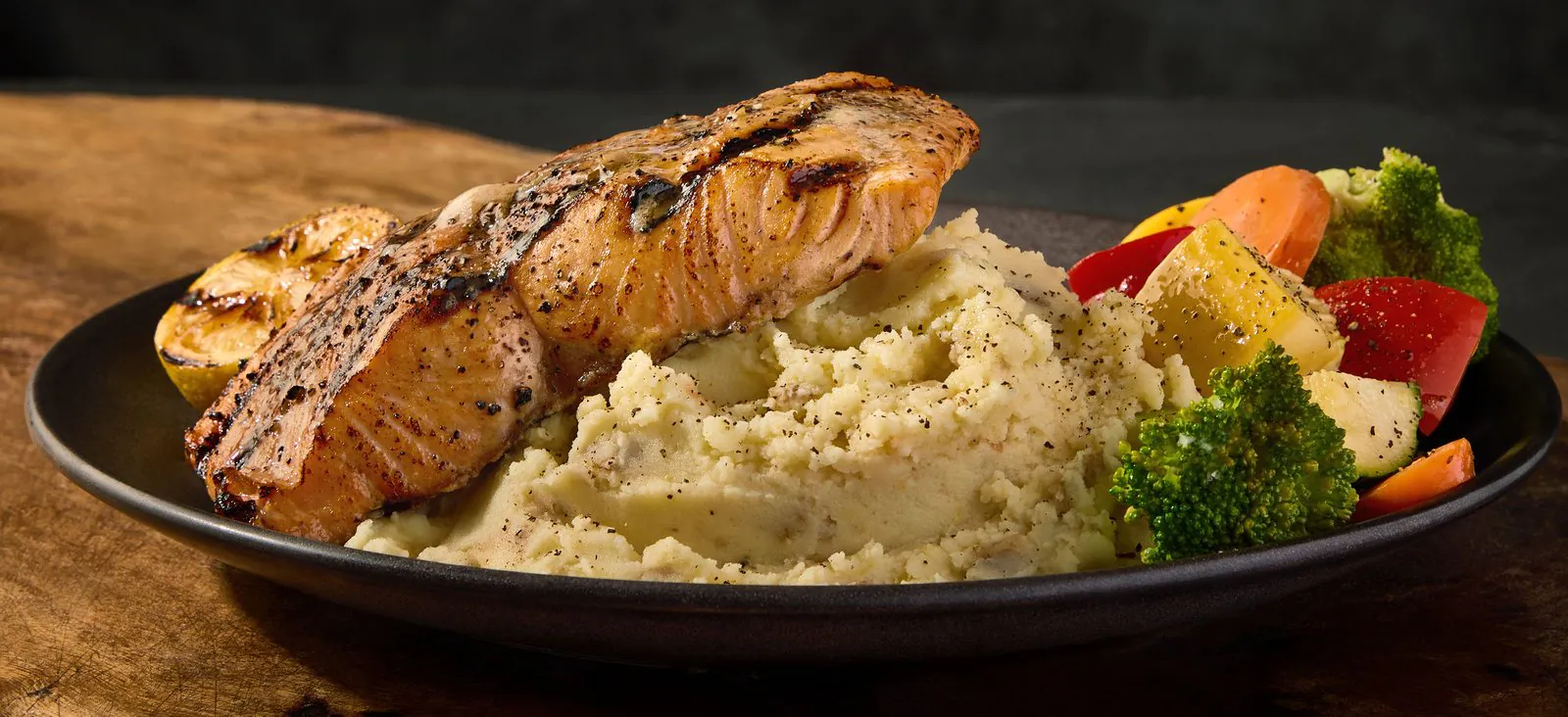 A wood-grilled salmon fillet with grill marks on a dark plate beside mashed potatoes, charred lemon and roasted vegetables