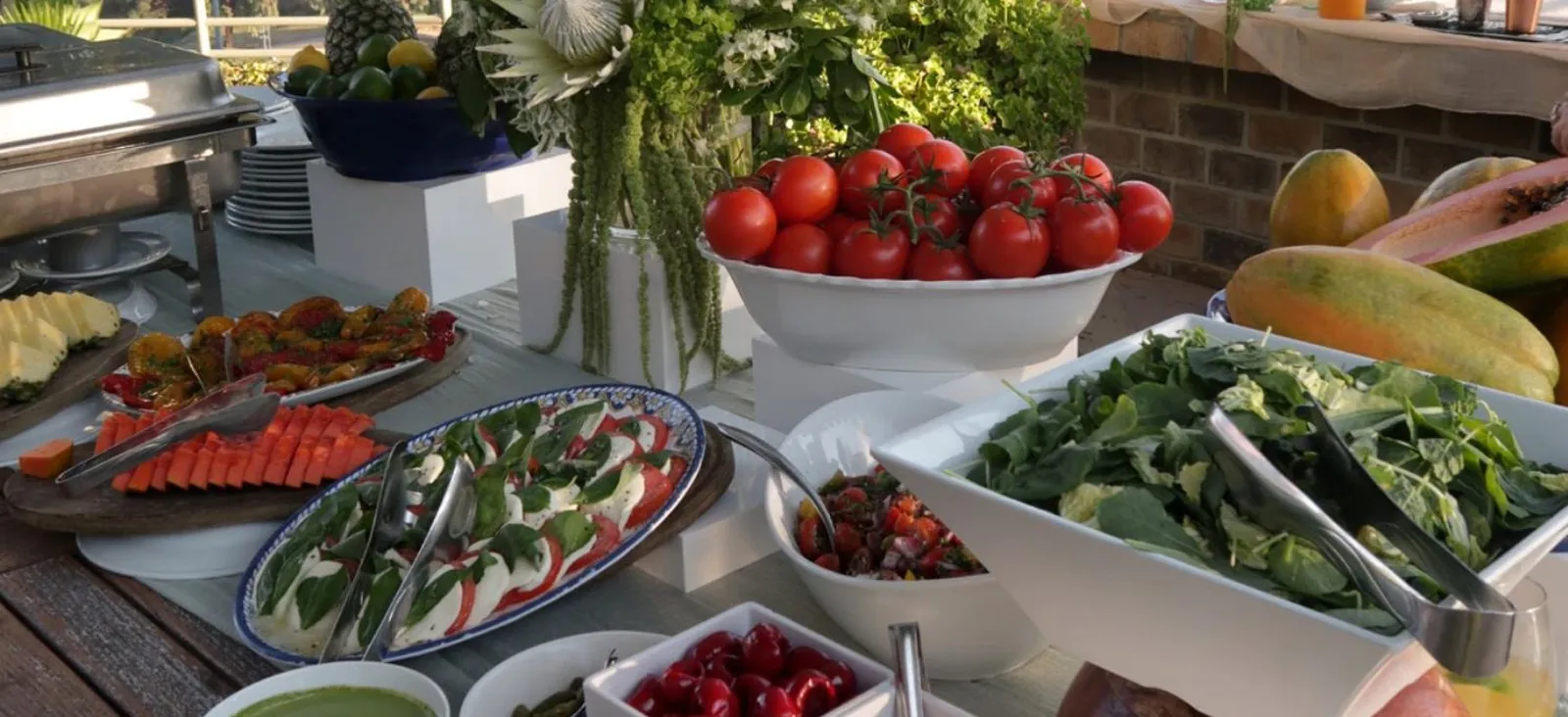 Outdoor catering table of separate salad trays, sliced fruit, whole tomatoes and greens beside a chafing dish
