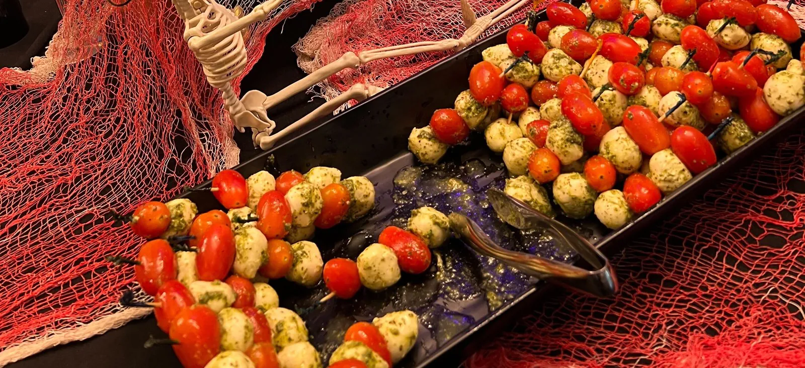 Caprese skewers on a black tray beside a toy skeleton and red cheesecloth at an office Halloween party