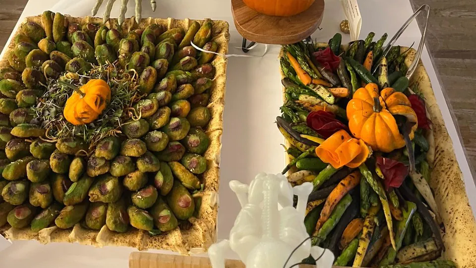 Two platters of roasted Brussels sprouts and roasted vegetables, each topped with a carved mini pumpkin, on a white buffet table