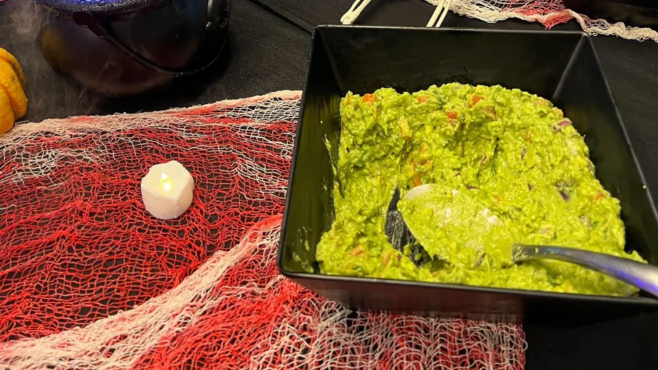 Black bowl of guacamole with a serving spoon on red cheesecloth beside a battery candle and a glowing cauldron prop