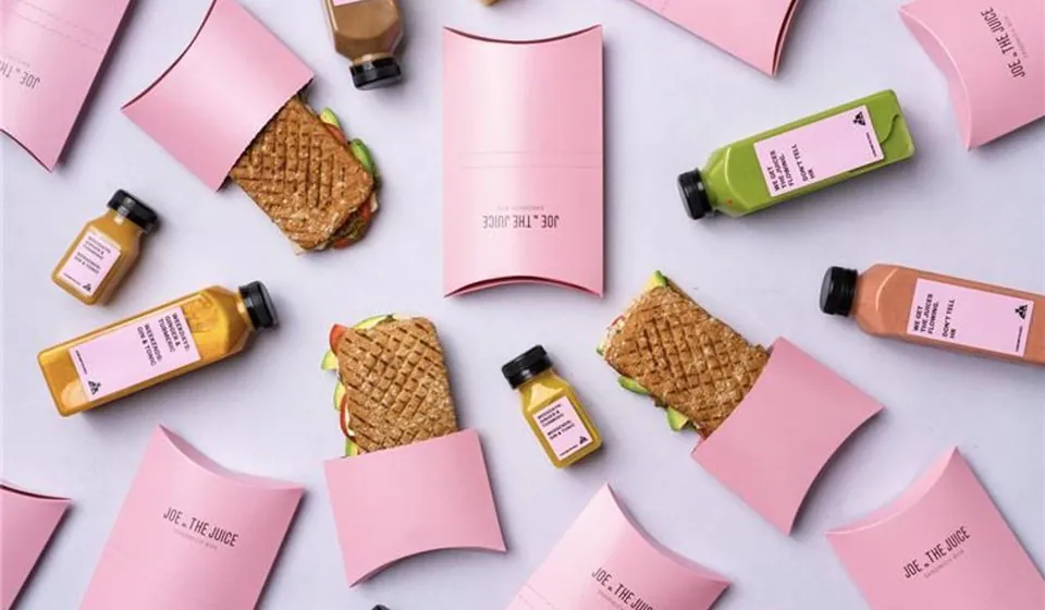 Toasted sandwiches, pink sandwich boxes and bottles of green and orange juice arranged on a pale surface