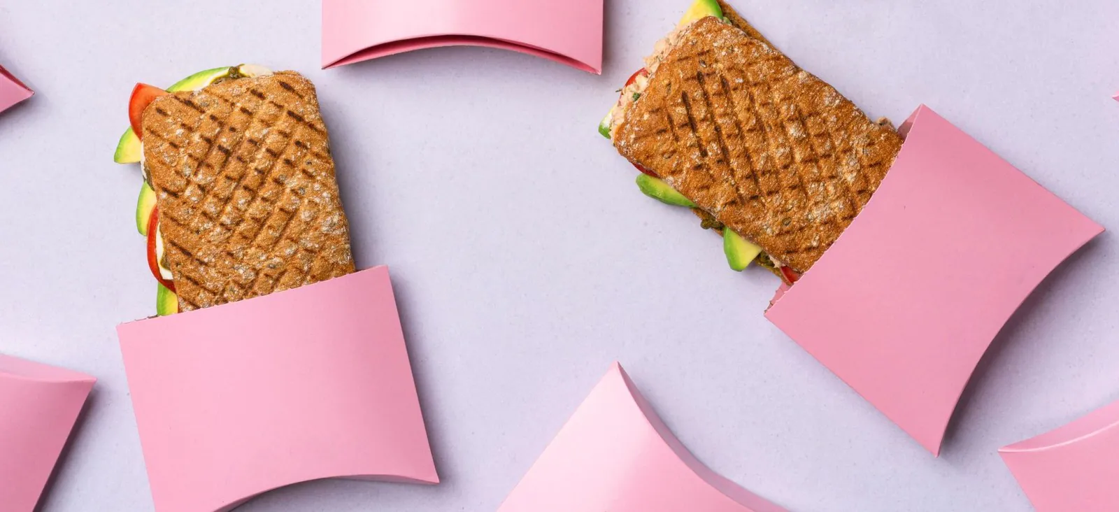 Two toasted catering sandwiches lying beside pale pink sandwich boxes on a light grey surface