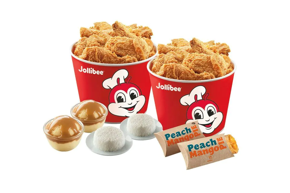 Two red Jollibee fried chicken buckets behind individual cups of gravy, two plated portions of steamed rice and two wrapped pies