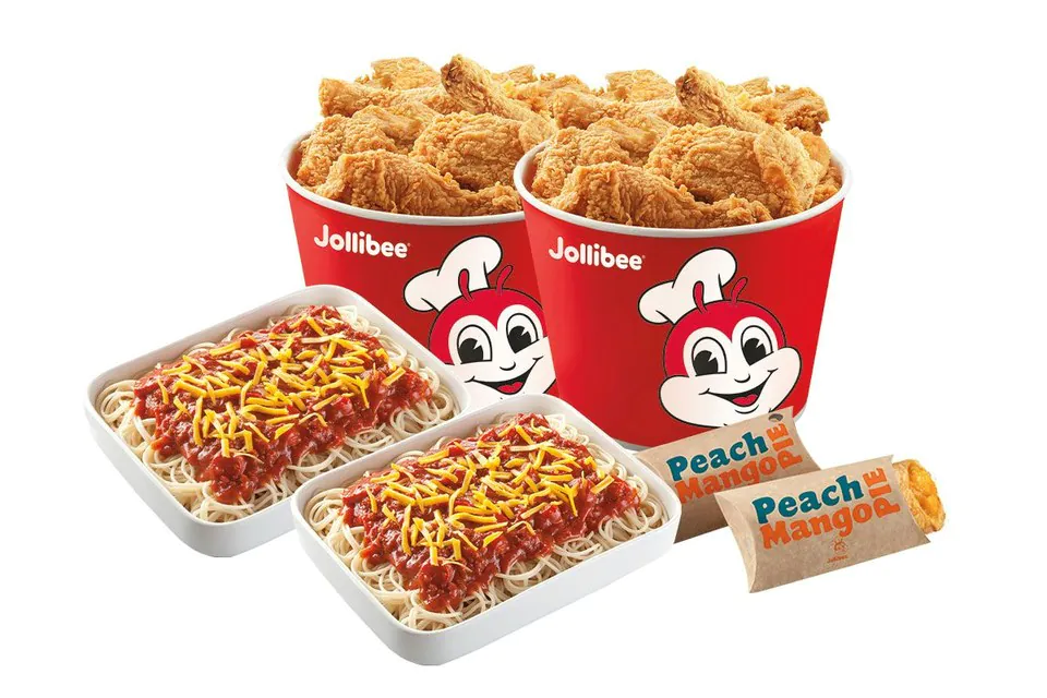 Two red Jollibee fried chicken buckets behind two white trays of spaghetti in tomato sauce topped with shredded cheese, and two wrapped pies