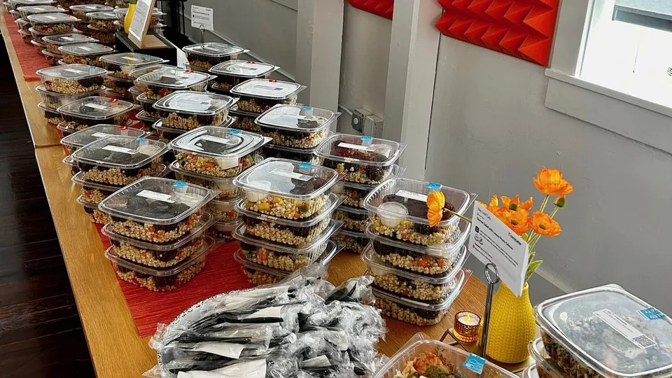 Long office table lined with lidded grain salads carrying printed item labels, a basket of wrapped cutlery and a vase of orange poppies