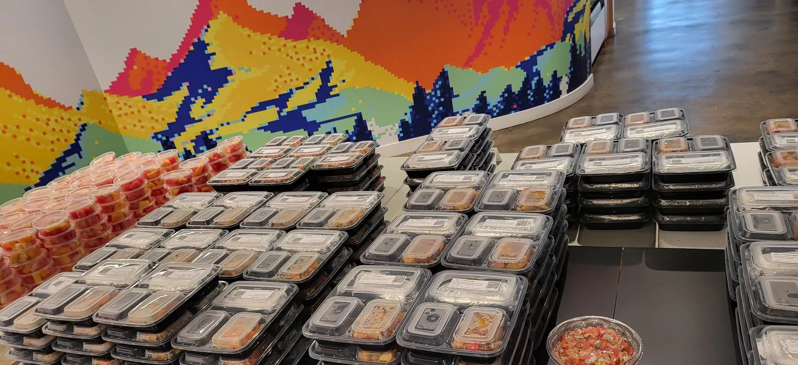 Rows of sealed, lidded compartment meals with printed item labels stacked on an office table beside cups of cut fruit