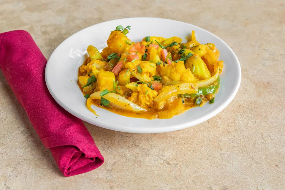 A white plate of aloo gobi, potato and cauliflower cooked with onion, tomato and green pepper, beside a rolled magenta napkin
