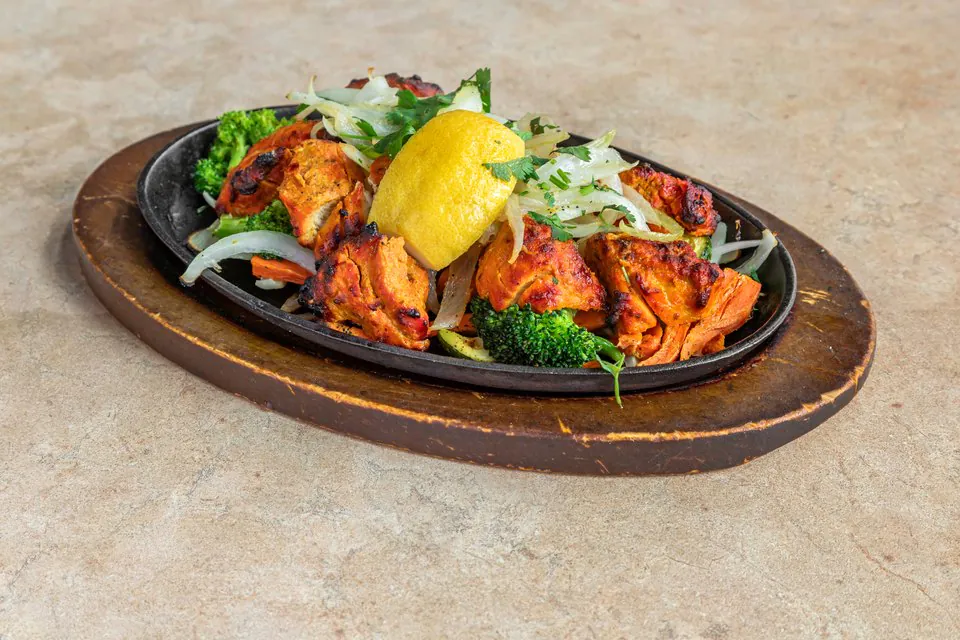 Chicken tikka on a cast iron sizzler platter with lemon, broccoli, carrot and sliced onion, set on a wooden board