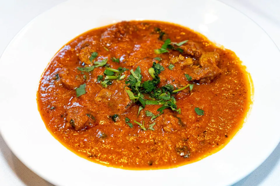 A white bowl of a tomato-based North Indian curry with tender pieces of meat, finished with chopped cilantro