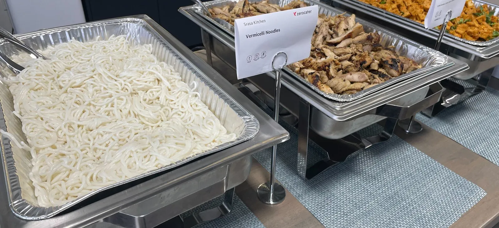 Foil catering pans of noodles and grilled chicken sitting in stainless chafing racks on an office kitchen counter