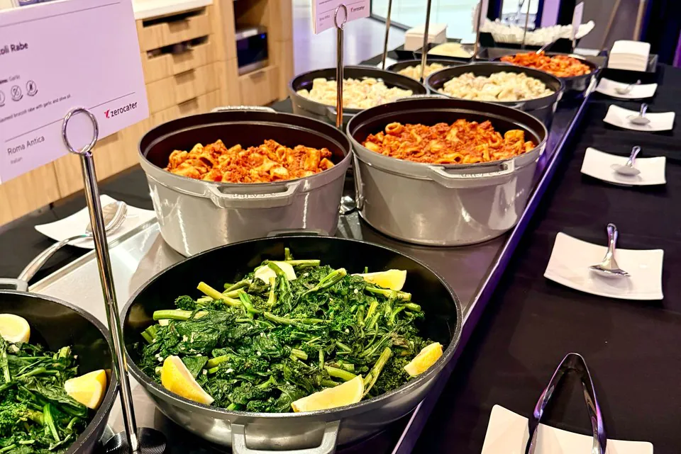 Red-sauce pasta in serving pans along an office buffet line with stacked plates and serving spoons