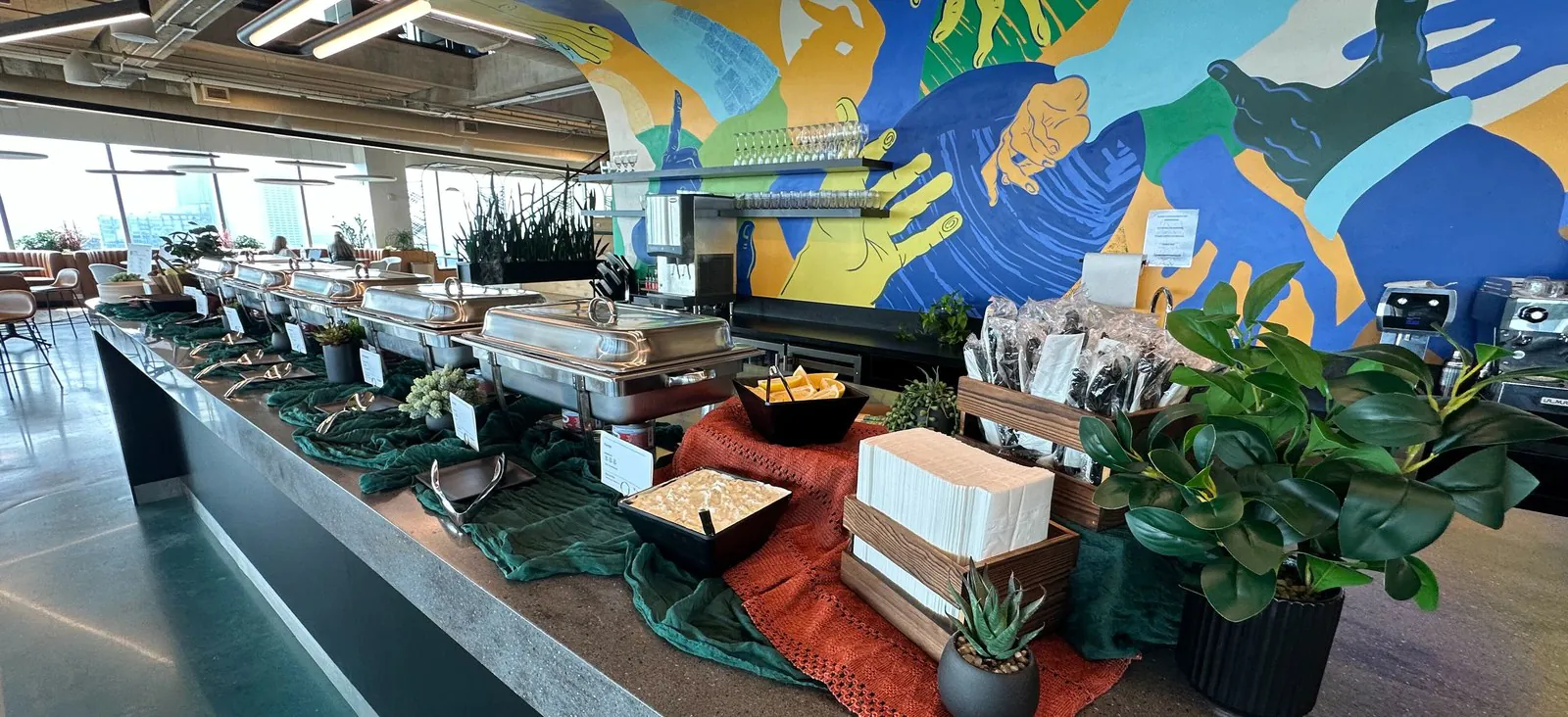 Long office counter holding a run of stainless chafing dishes with serving spoons, a napkin caddy and wrapped cutlery, under a painted mural