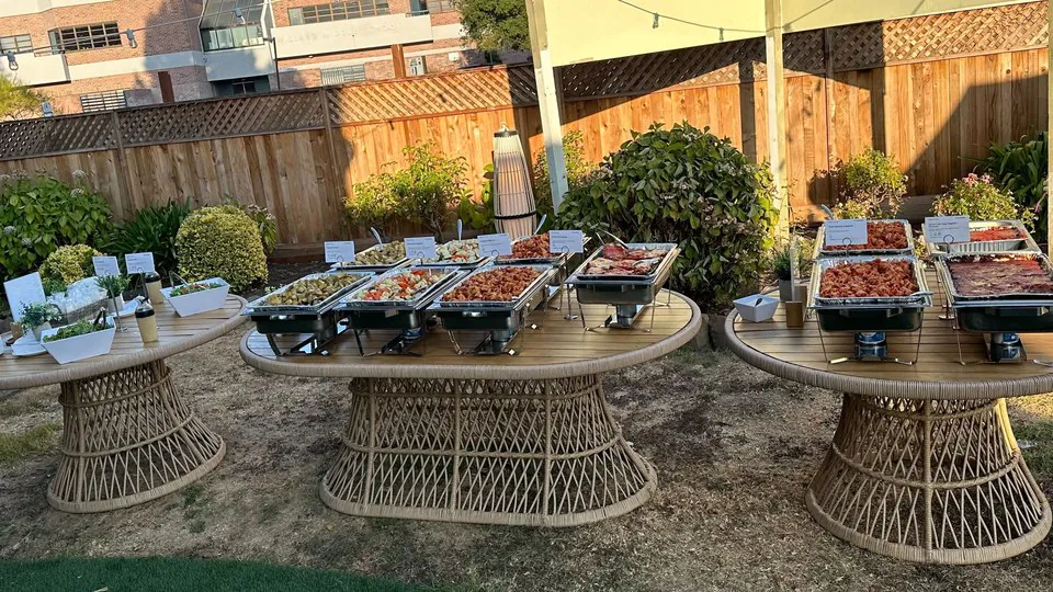 Three rattan tables in a sunlit backyard, each holding stainless chafing pans over burners with printed dish cards