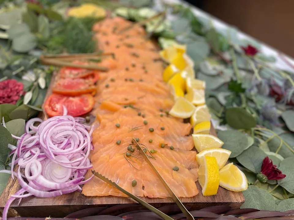 Long wooden board of sliced smoked salmon with capers, lemon wedges, red onion and tomato on a table dressed with eucalyptus