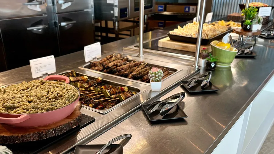 Stainless steel serving line in an office with trays of skewers, dips and sliced bread behind a sneeze guard