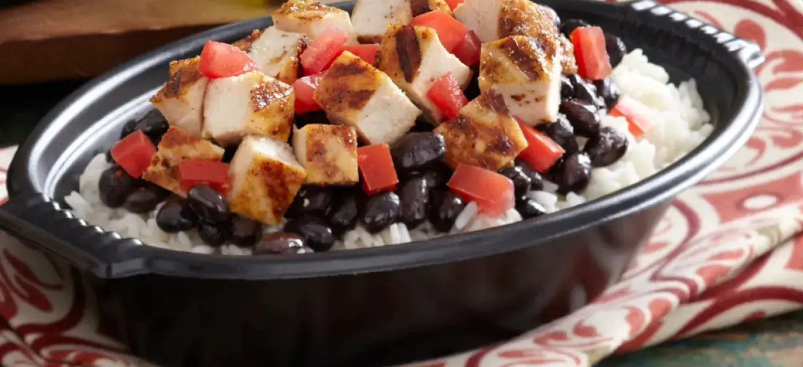 A black catering bowl of white rice and black beans topped with cubes of grilled chicken breast and diced tomato, on a red patterned napkin