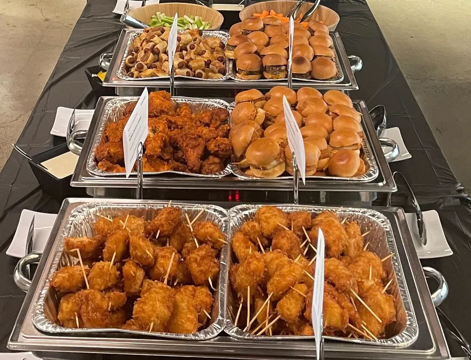 A long office buffet line of foil trays holding small burgers on buns and fried bites, with serving tongs on white plates