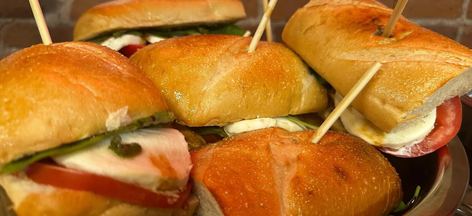 Sandwiches on toasted rolls held together with wooden picks, stacked on a steel catering tray against a brick wall