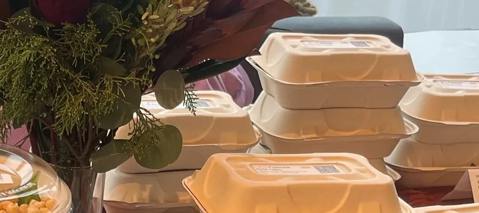 Stacks of sealed fibre clamshell boxes on an office table beside lidded bowls and a floral arrangement