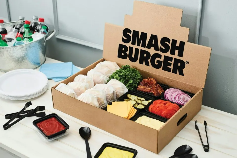 An open Smashburger catering box holding individually wrapped burgers beside a compartment tray of sliced cheese, tomatoes, red onion, pickles, bacon and sauces