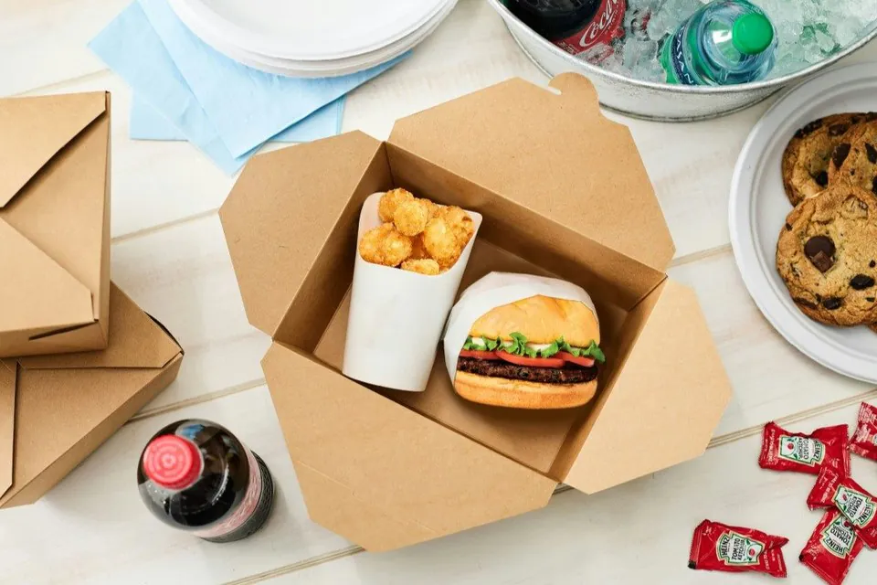 A kraft boxed lunch open on a table showing a cheeseburger and a cup of tots, with sealed ketchup sachets and a bottled soda beside it