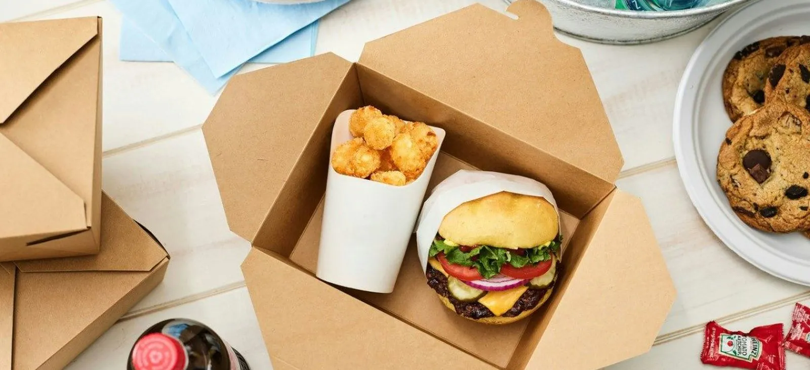 An open kraft catering box holding a cheeseburger and a cup of tots, with stacked spare boxes, cookies on a plate and loose ketchup sachets on a white table
