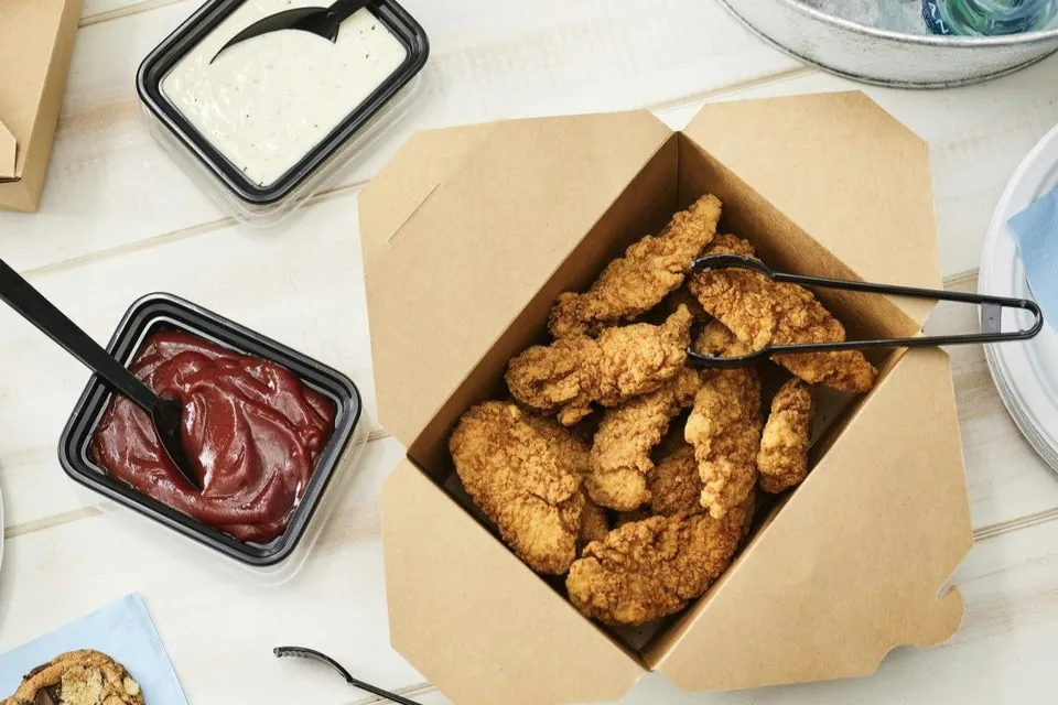 An open catering box of crispy chicken tenders with serving tongs, beside two sealed tubs of dipping sauce