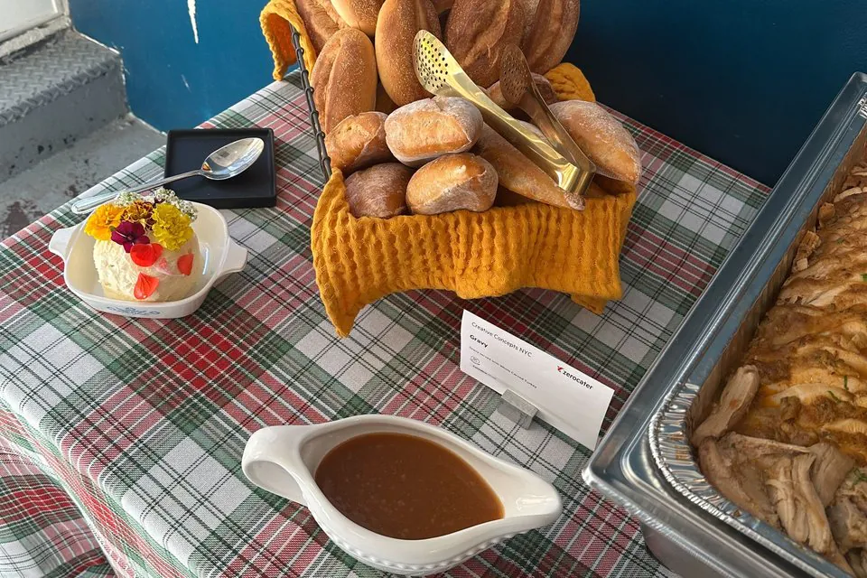White gravy boat on a plaid tablecloth beside a basket of dinner rolls and a tray of carved turkey at an office Thanksgiving lunch