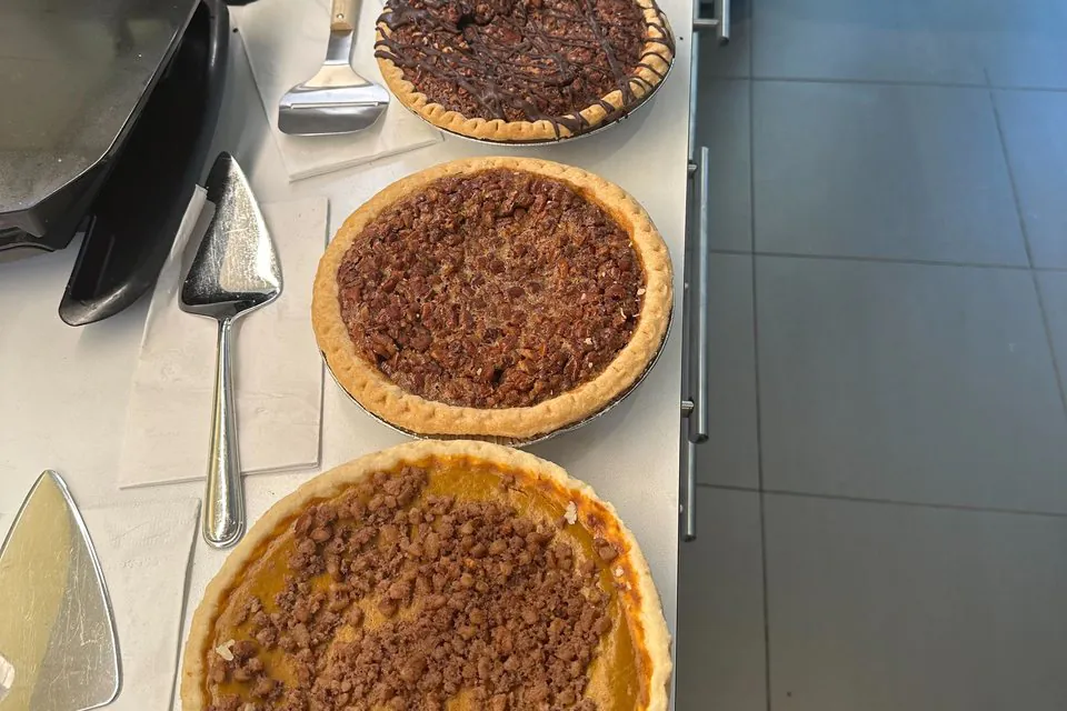Whole pecan and pumpkin pies with a chocolate-drizzled pie on a stainless office counter beside two pie servers