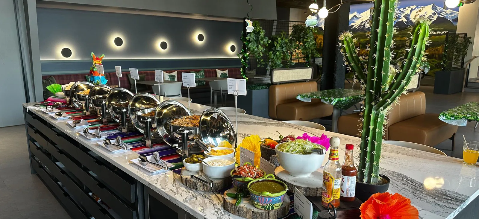 An office taco buffet island with a row of chafing dishes, bowls of salsa and guacamole on wood risers, and printed menu cards along a marble counter