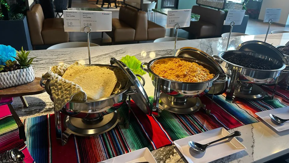 Three chafing dishes holding warm tortillas, Mexican rice and black beans on a striped runner, each with a printed card naming the dish