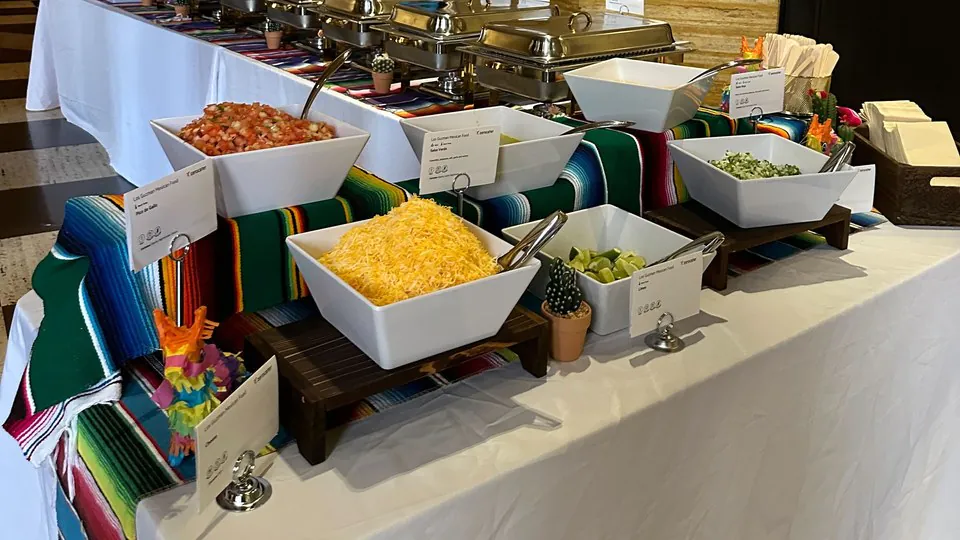 A long taco bar with white bowls of pico de gallo, shredded cheese, limes and chopped onion on wood risers, each bowl with a printed card beside it