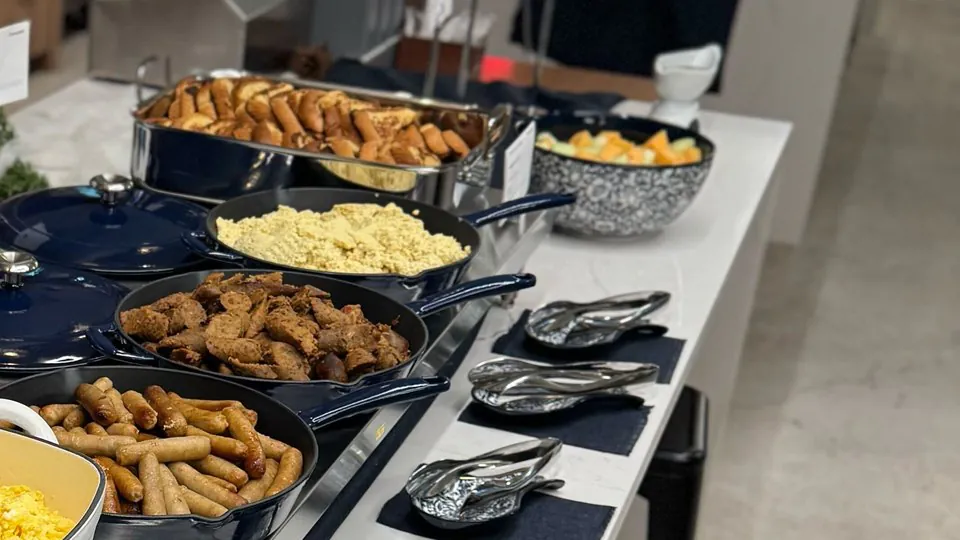 Catering pans of scrambled egg, sausage links, seasoned sausage and breakfast pastries lined up along an office counter with serving spoons