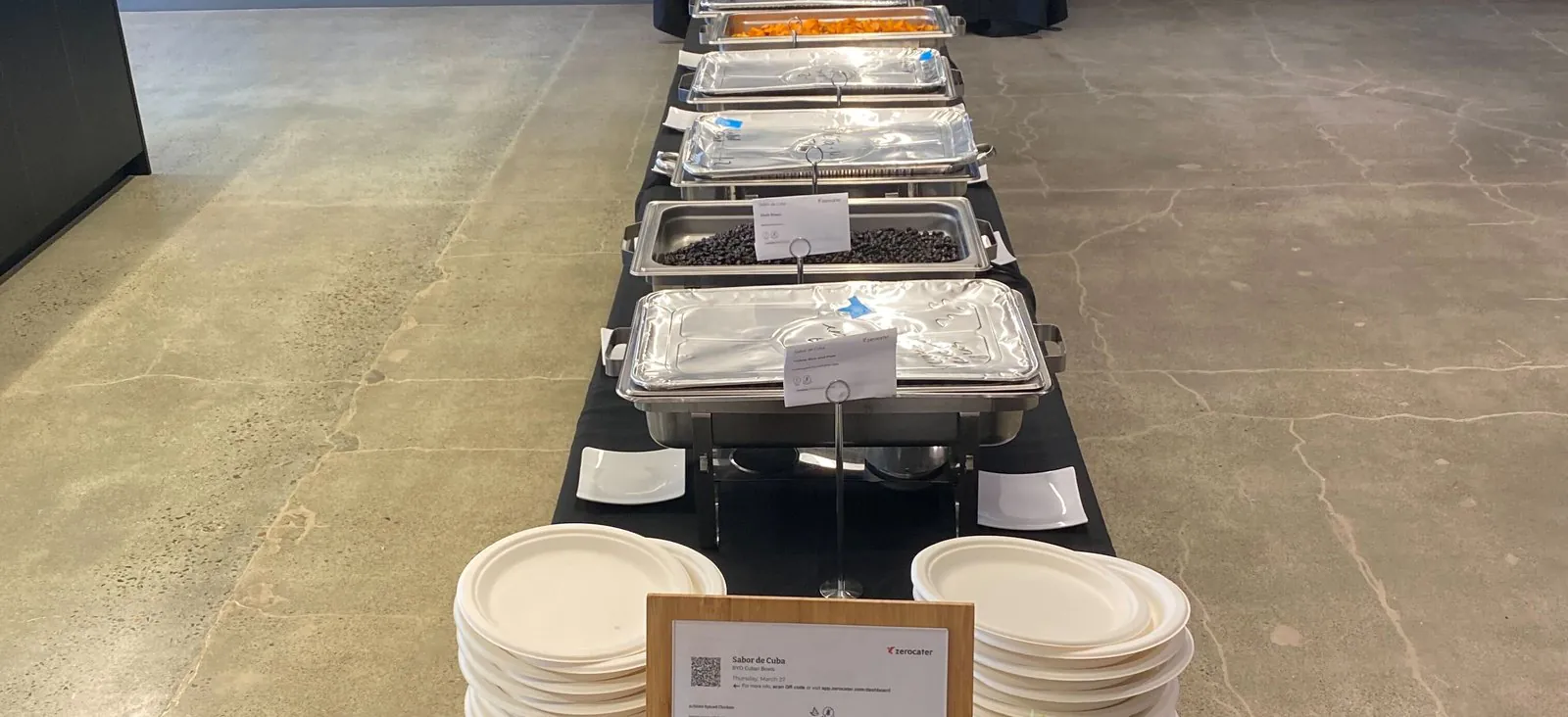 A long office buffet line of lidded foil chafing trays down a black-draped table, with stacked plates and a printed menu card at the near end