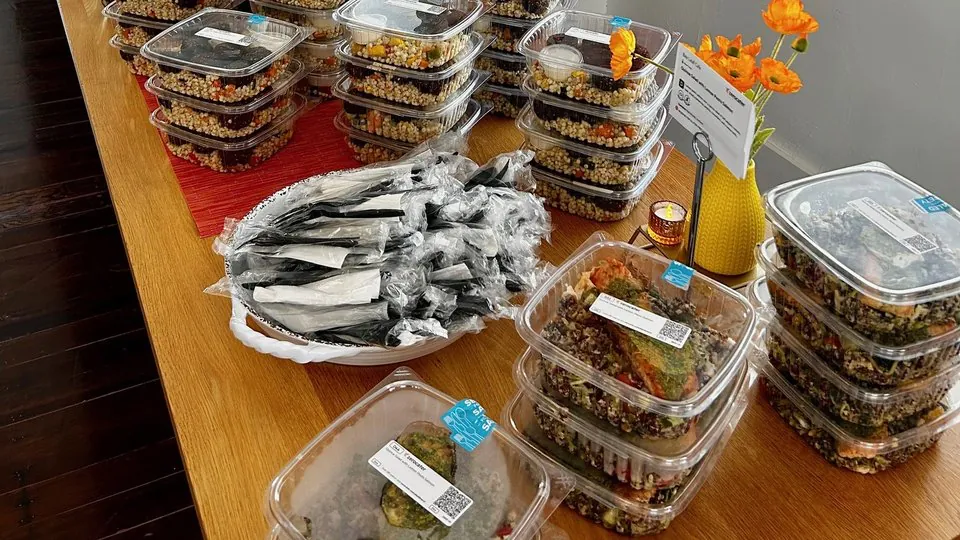 Rows of sealed single-serve meal containers stacked on a wooden table beside a large pile of individually wrapped cutlery sets