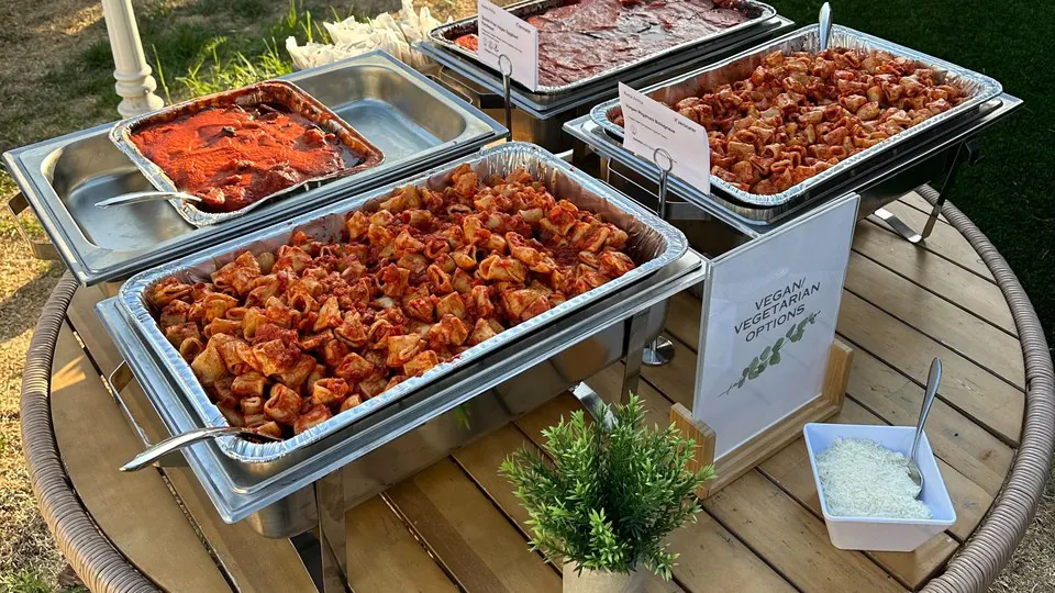 Outdoor work event with chafing pans of pasta and a printed card reading Vegan Vegetarian Options standing beside them