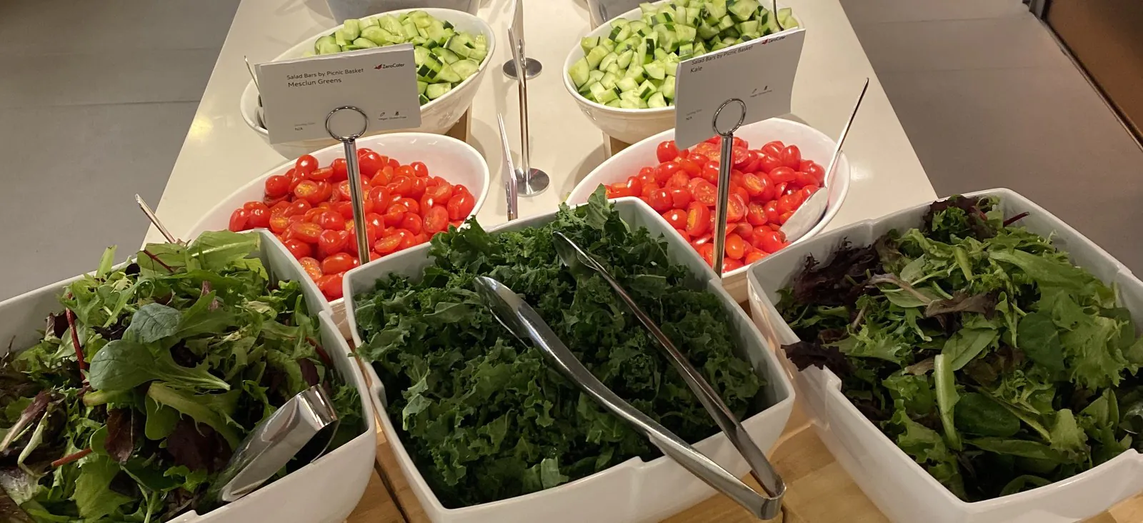 Office salad bar with mesclun greens, kale, cherry tomatoes and cucumber each in a separate white bowl behind printed cards naming the component