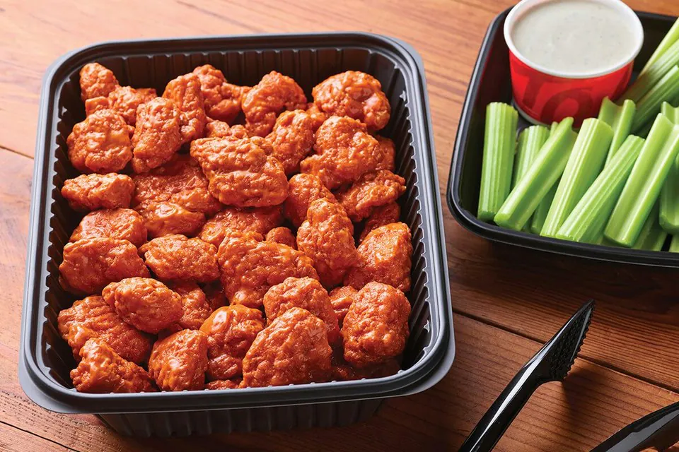 A black catering tray of Applebee's boneless wings in a red sauce beside a tray of celery sticks and a cup of blue cheese dressing on a wood table