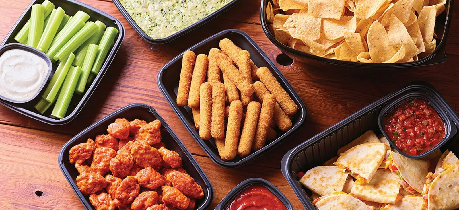 Applebee's Classic Combo catering spread on a wood table: boneless wings, mozzarella sticks, spinach and artichoke dip, chicken quesadilla wedges, celery with ranch and a bowl of tortilla chips