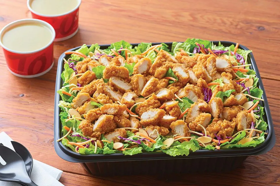 An Applebee's catering tray of Oriental Chicken Salad with crispy chicken tenders, greens, crunchy noodles and almonds, beside two cups of vinaigrette