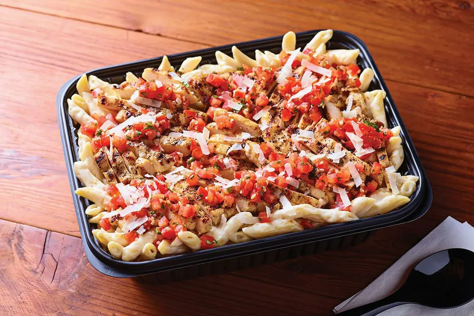 An Applebee's catering tray of Three-Cheese Chicken Penne topped with grilled chicken, diced bruschetta tomatoes and shaved Parmesan on a wood table